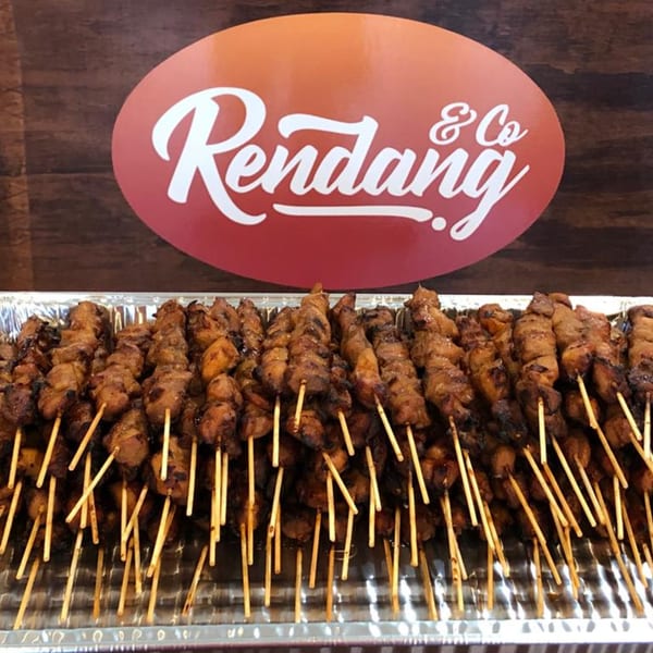 Numerous Indonesian chicken satay skewers in a foil tray, with the Rendang & Co logo in the background.