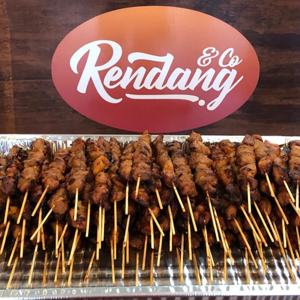 Numerous Indonesian chicken satay skewers in a foil tray, with the Rendang & Co logo in the background.