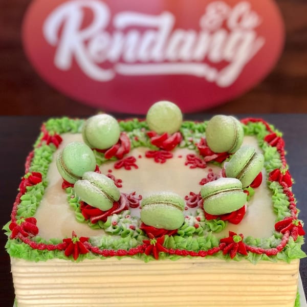 Square cake with green and red frosting, topped with green macarons, in front of a blurred Rendang & Co. sign.