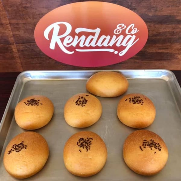 Seven golden-brown buns with chocolate sprinkles on a metal tray, with the Rendang & Co logo in the background.