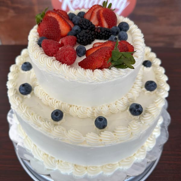 Two-tiered white cake with piped frosting, topped with strawberries, blueberries, raspberries, and blackberries.