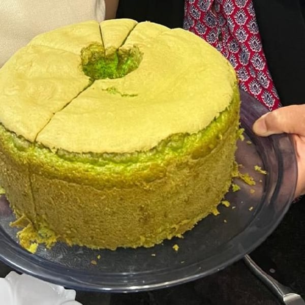 A large, round pandan chiffon cake with a hole in the center, partially sliced, on a clear plate.