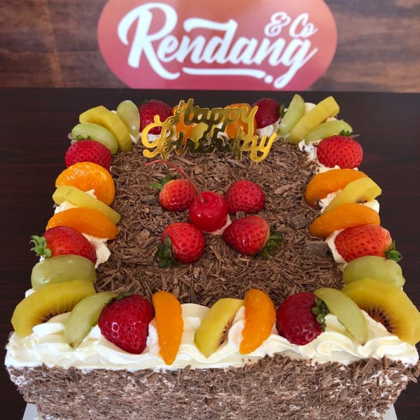 Square chocolate cake with whipped cream, fresh fruit, and a "Happy Birthday" topper.