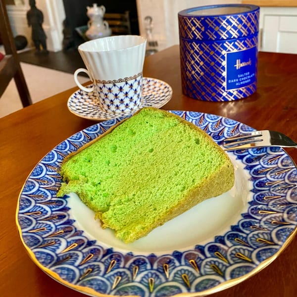 A slice of vibrant green pandan cake on a blue and white patterned plate with a teacup and Harrods tin.