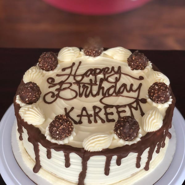 White frosted birthday cake with chocolate drizzle, Ferrero Rocher candies, and "Happy Birthday KAREEM" in chocolate script.