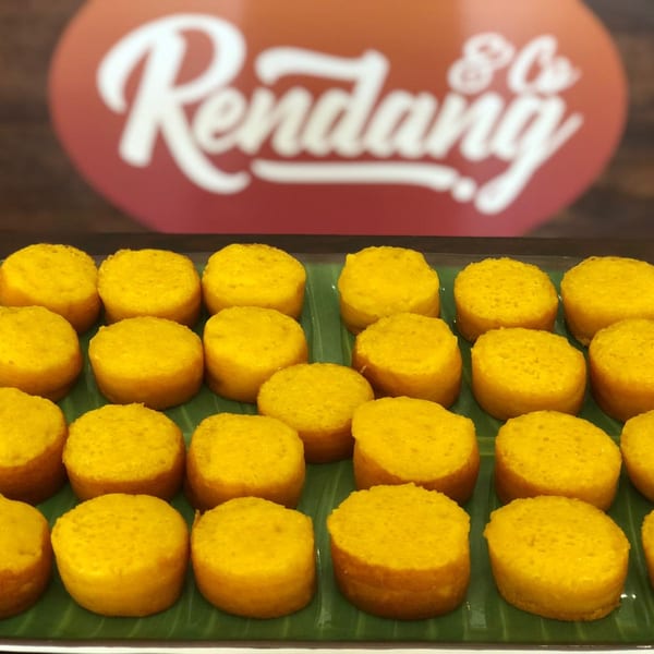 Rows of small, round, yellow Indonesian cakes on green banana leaves with the Rendang & Co. logo in the background.