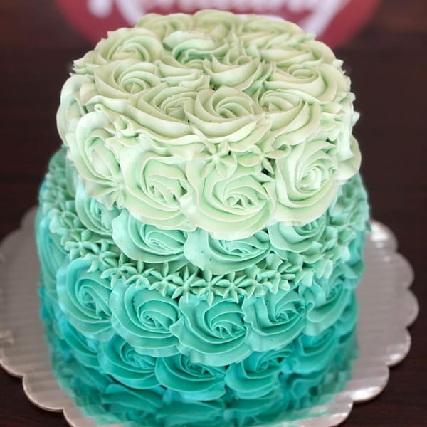 Two-tiered cake with ombre teal and light green frosting roses on a silver scalloped plate.