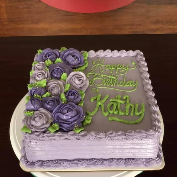 Square birthday cake with purple and lavender roses, green leaves, and "Happy Birthday Kathy" in green icing.
