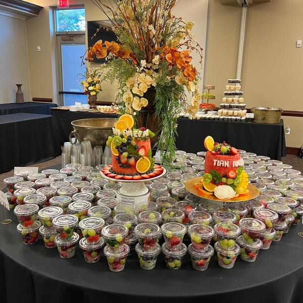 Two fruit-decorated cakes and numerous fruit cups on a black-draped table with floral arrangements.