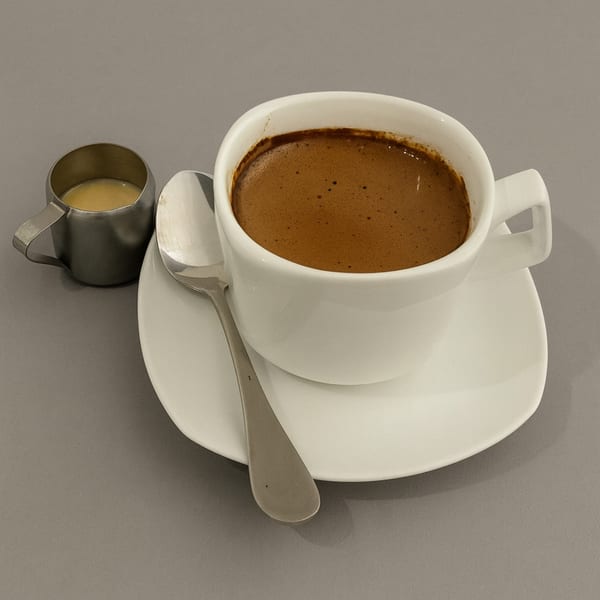 A white cup of coffee with a spoon and a small metal creamer on a gray surface.