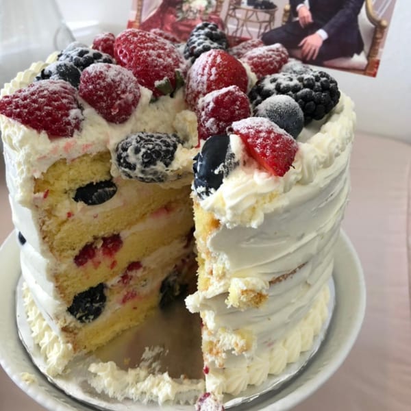 A partially sliced vanilla cake with white frosting, topped with raspberries, blackberries, and blueberries.