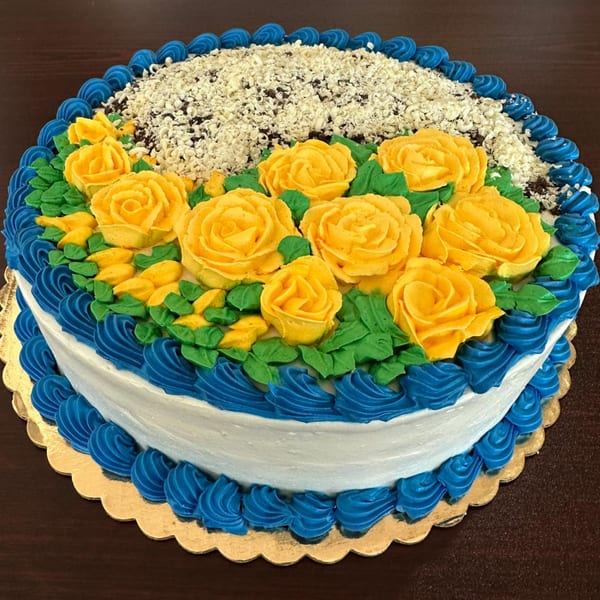 A round cake with white frosting, blue piped borders, yellow roses, green leaves, and white chocolate shavings.