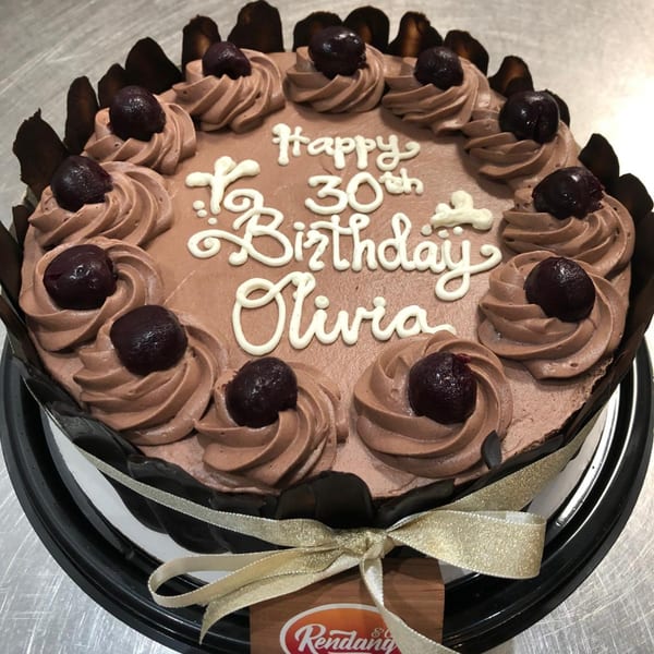 Chocolate birthday cake with "Happy 30th Birthday Olivia" in white icing, cherries, and chocolate swirls.