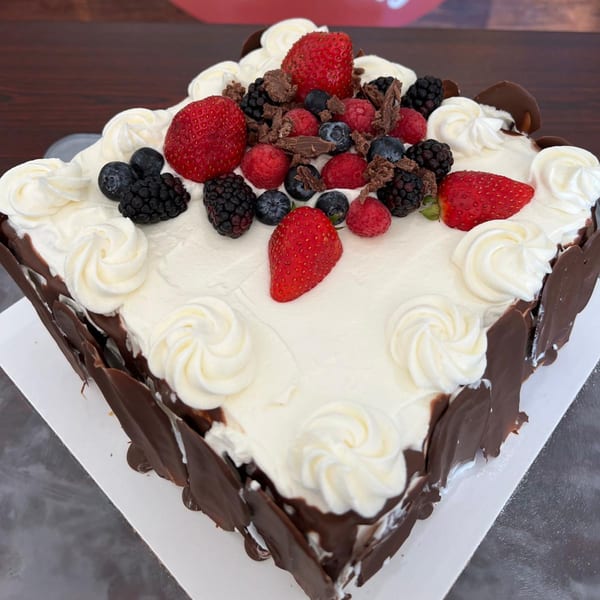 Square cake with white frosting, chocolate shards, and a variety of fresh berries on top.
