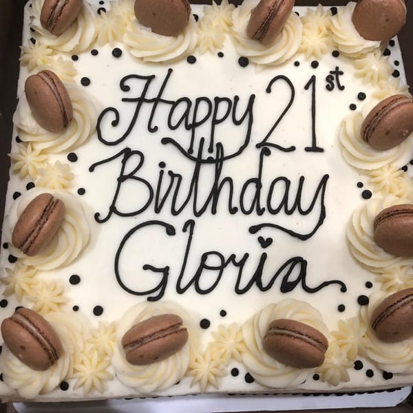White birthday cake with "Happy 21st Birthday Gloria" in black icing, decorated with macarons.