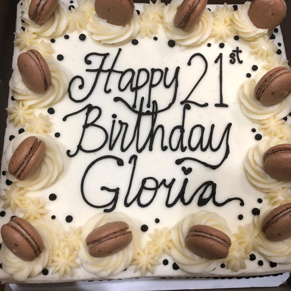 White birthday cake with "Happy 21st Birthday Gloria" in black icing, decorated with macarons.